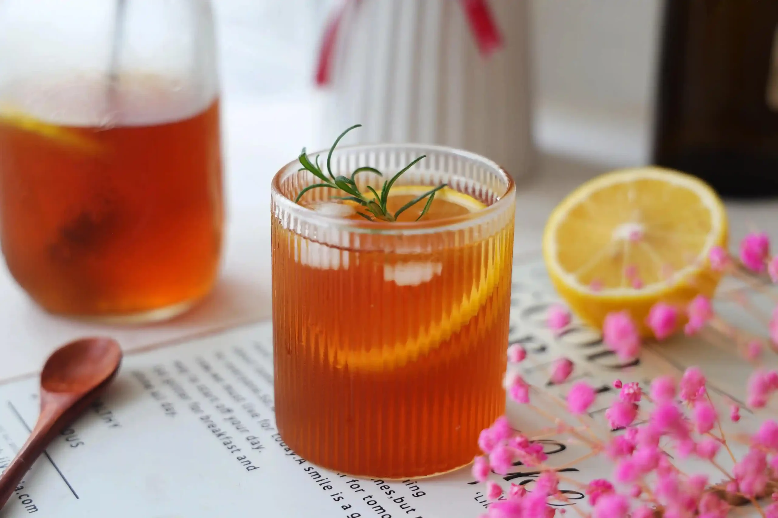 A visually appealing glass of iced black tea, garnished with lemon and rosemary, complemented by pink flowers and a lemon slice for a refreshing look.