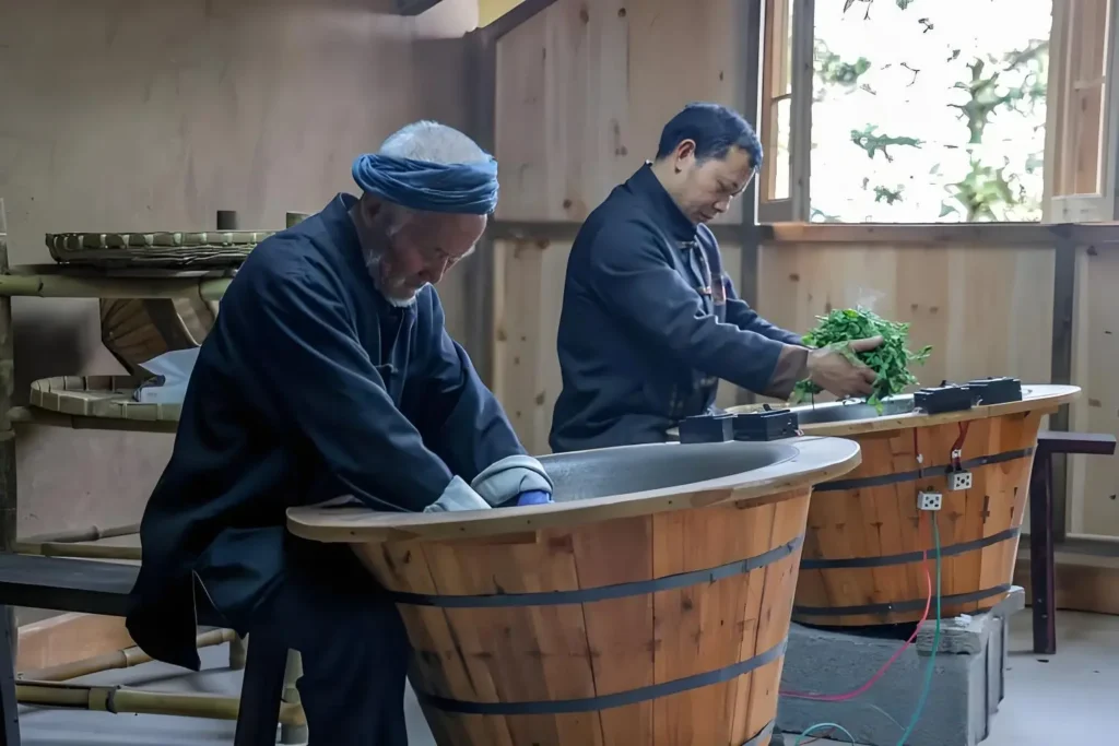 Two individuals engaged in traditional Chinese tea processing, reflecting the rich heritage and origin of Chinese tea. They work with wooden utensils, surrounded by a rustic setting with bamboo racks and natural light, emphasizing the artisanal craftsmanship involved in tea making.